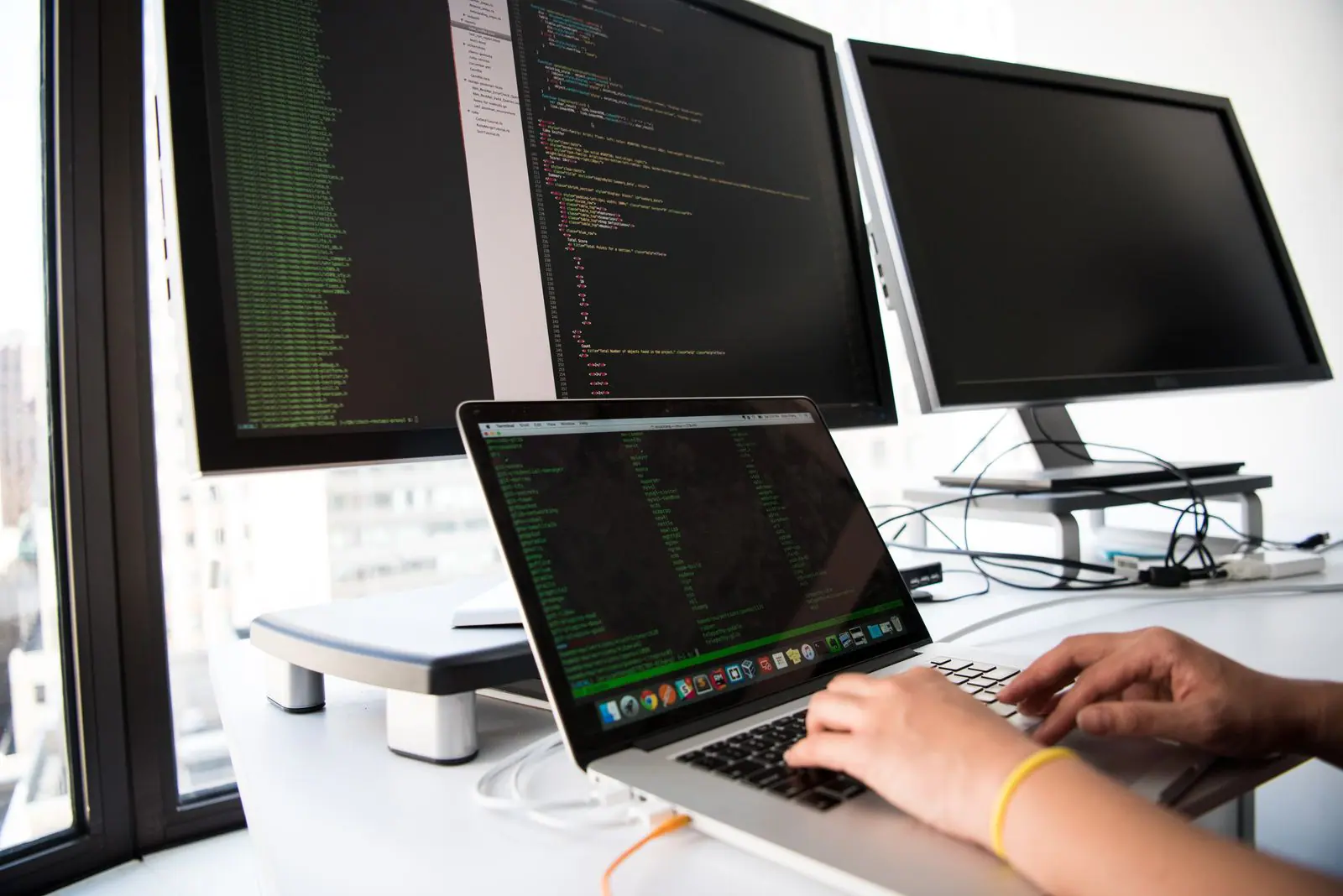 Developer typing on a laptop in front of two monitors of code